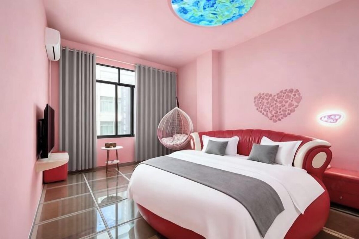 Habitación de hotel en tonos rosados con una cama grande, una silla colgante, una mesita pequeña y una decoración de corazón en la pared.