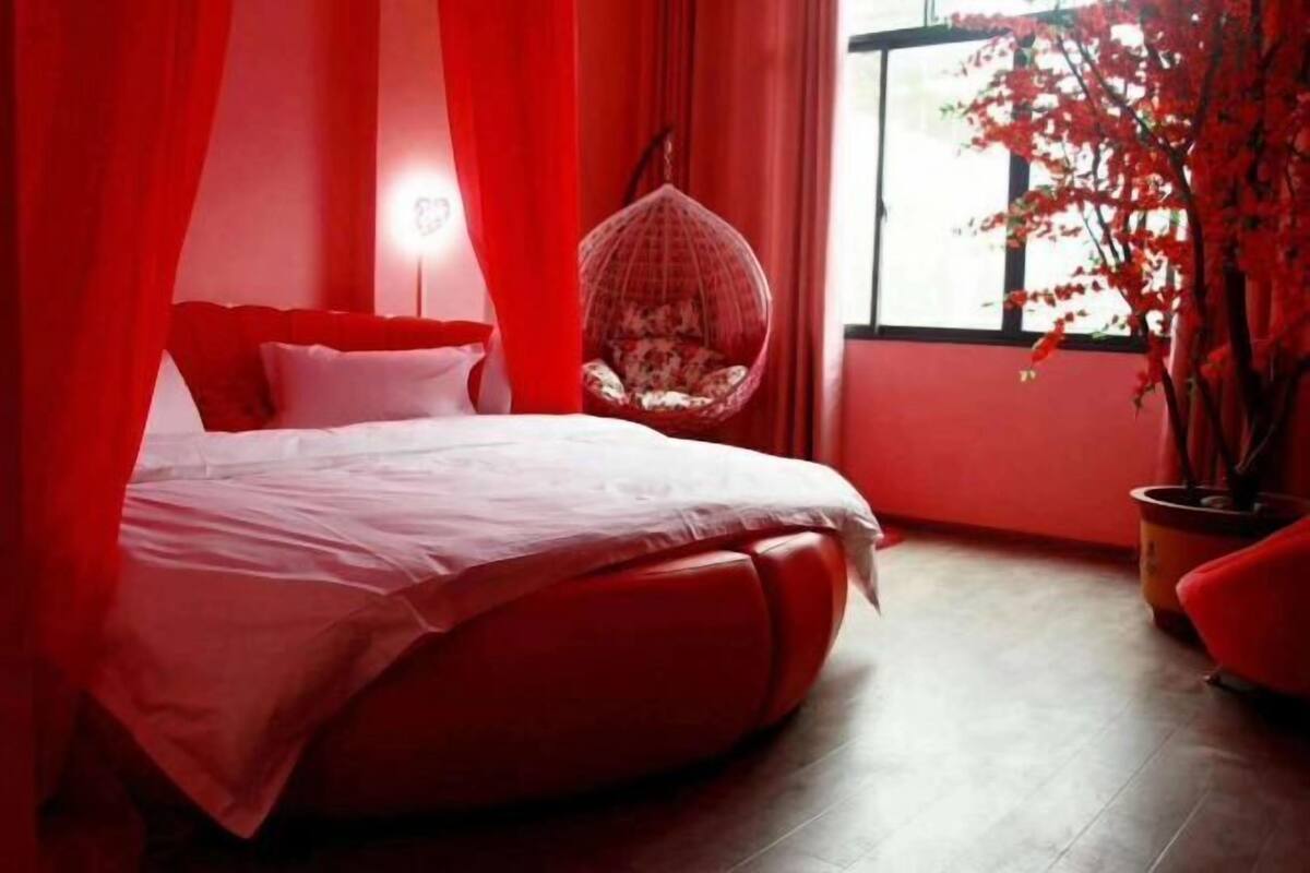 A bedroom with a bed, red curtains, a hanging chair, and a potted plant.