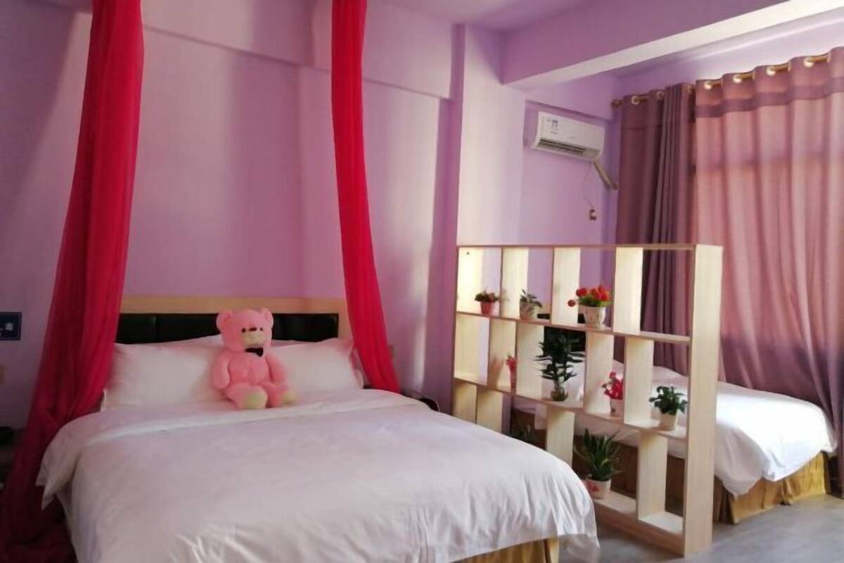 A bedroom with a bed, a pink teddy bear, a wooden shelf with plants, and a window with curtains.