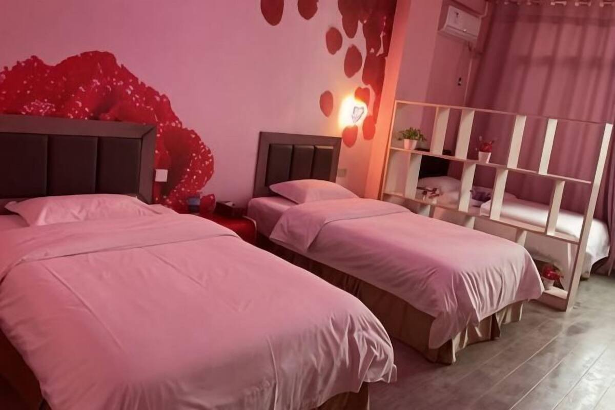 A room with two beds, a red decorative wall, a white shelf unit, and a pink curtain.
