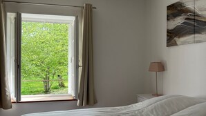 A bedroom with a large bed, a window with curtains, a bedside table with a lamp, and a painting on the wall.