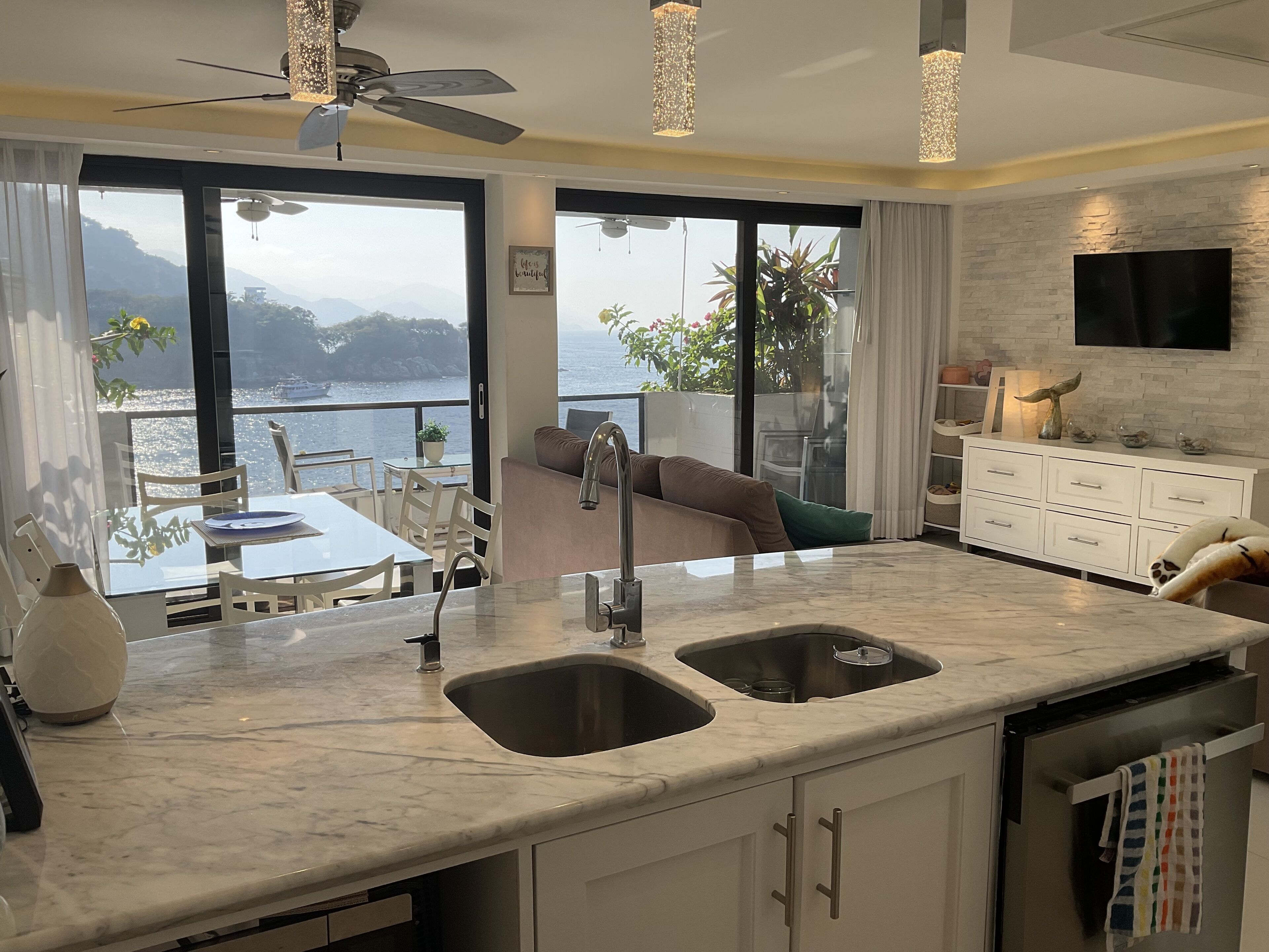 A modern kitchen with a large island, a dining area with a view, and a living area with a sofa and TV.