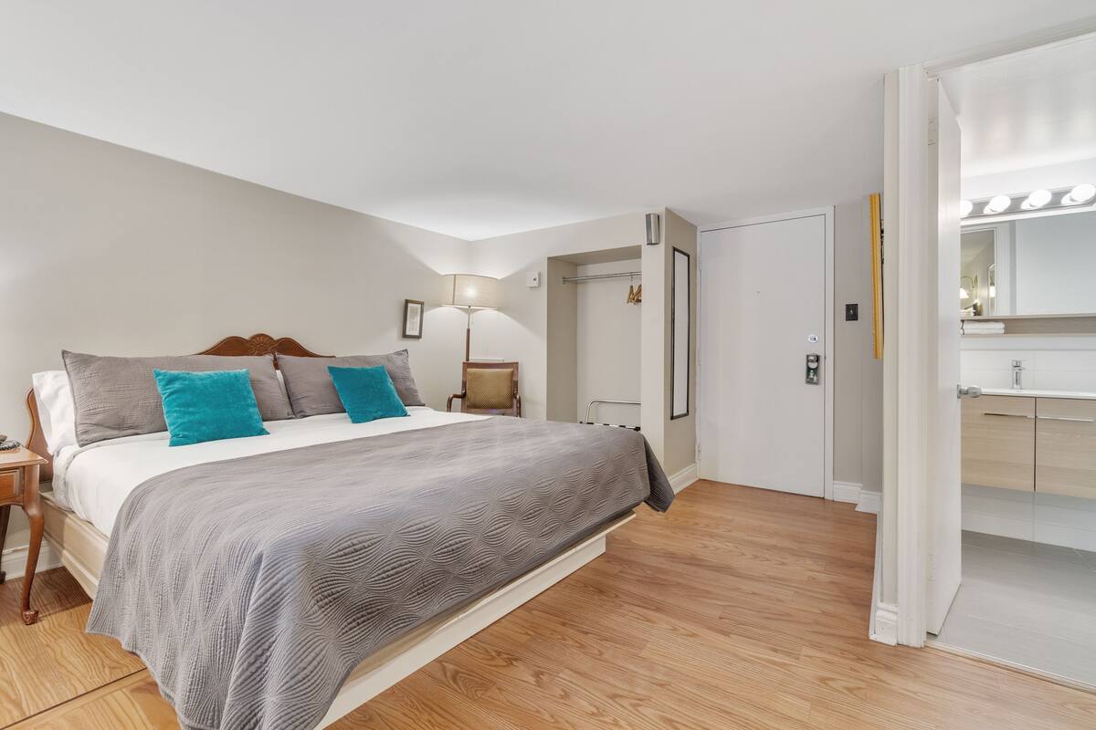 Une chambre à coucher comprenant un lit, une table de chevet, une lampe, une porte donnant sur une salle de bain et un miroir avec éclairage.
