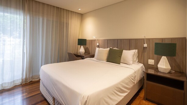Habitación de hotel con una cama grande, dos mesitas de noche con lámparas, una silla y vistas al exterior a través de cortinas traslúcidas.