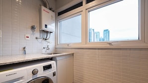 A laundry room with a Samsung washing machine, a Rinnai water heater, and a window with a city view. - Edifício Triplo (São Paulo)