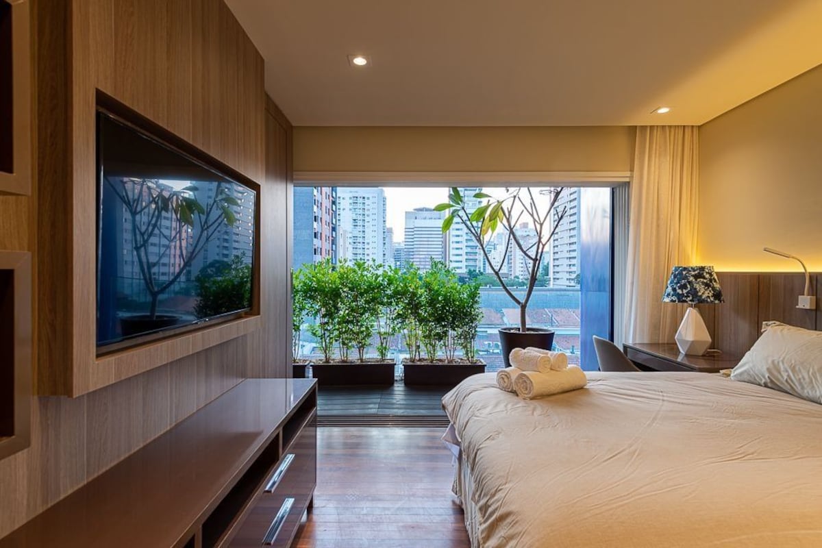 A modern hotel room with a large bed, a flat-screen TV mounted on the wall, a desk with a lamp, and a view of the city through large windows.