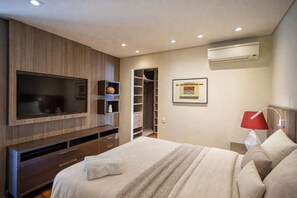 A modern bedroom with a large TV, built-in shelving, a walk-in closet, and an air conditioning unit. - Edifício Triplo (São Paulo)