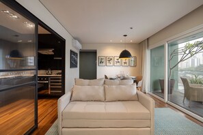 A modern living room with a sofa, a dining area with a table and chairs, and a kitchen visible through a glass door. - Edifício Triplo (São Paulo)