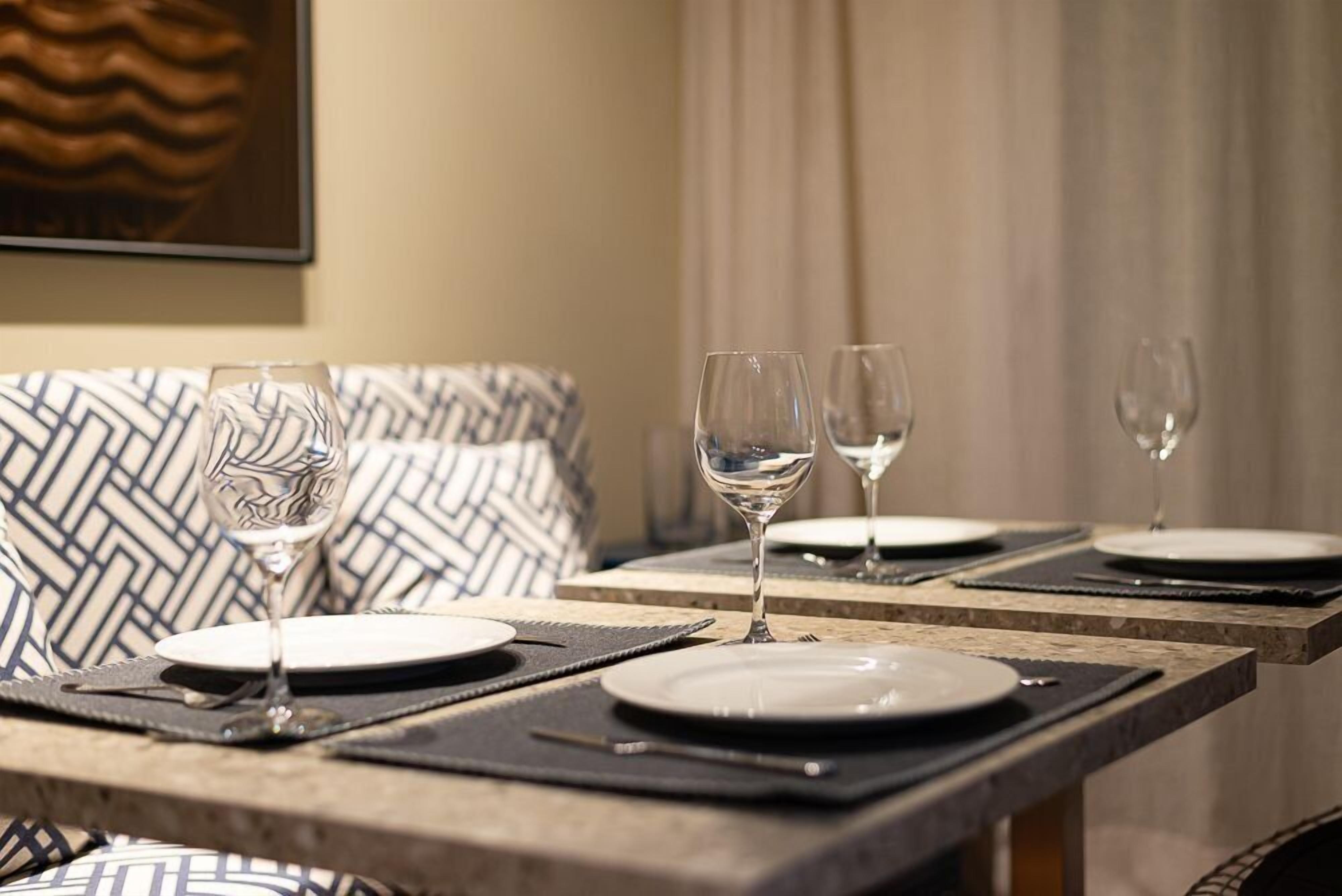 A dining table set with plates, cutlery, and wine glasses, with a patterned cushion in the background.
