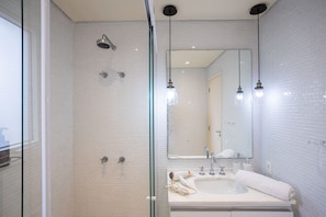 A modern bathroom with white brick walls, a glass shower enclosure, a sink with a towel, and pendant lighting. - Edifício Triplo (São Paulo)