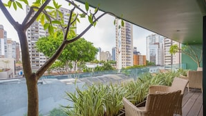 A balcony with wicker furniture, potted plants, and a view of a cityscape and river. - Edifício Triplo (São Paulo)