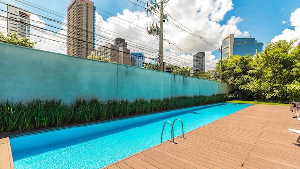 A swimming pool with a wooden deck, surrounded by a high wall and greenery, with city buildings in the background. - Edifício Triplo (São Paulo)