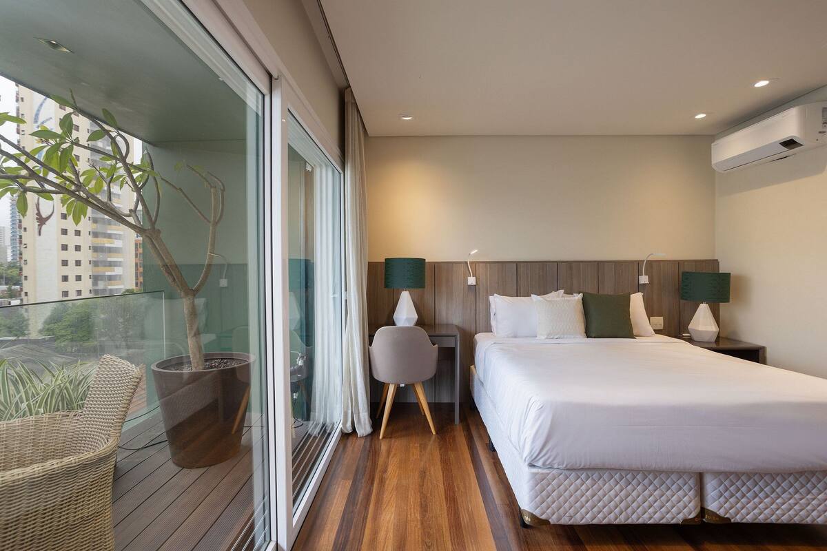 Ein modernes Hotelzimmer mit einem großen Bett, einer Zimmerpflanze, einem Korbstuhl und Blick auf die Stadt durch raumhohe Fenster.