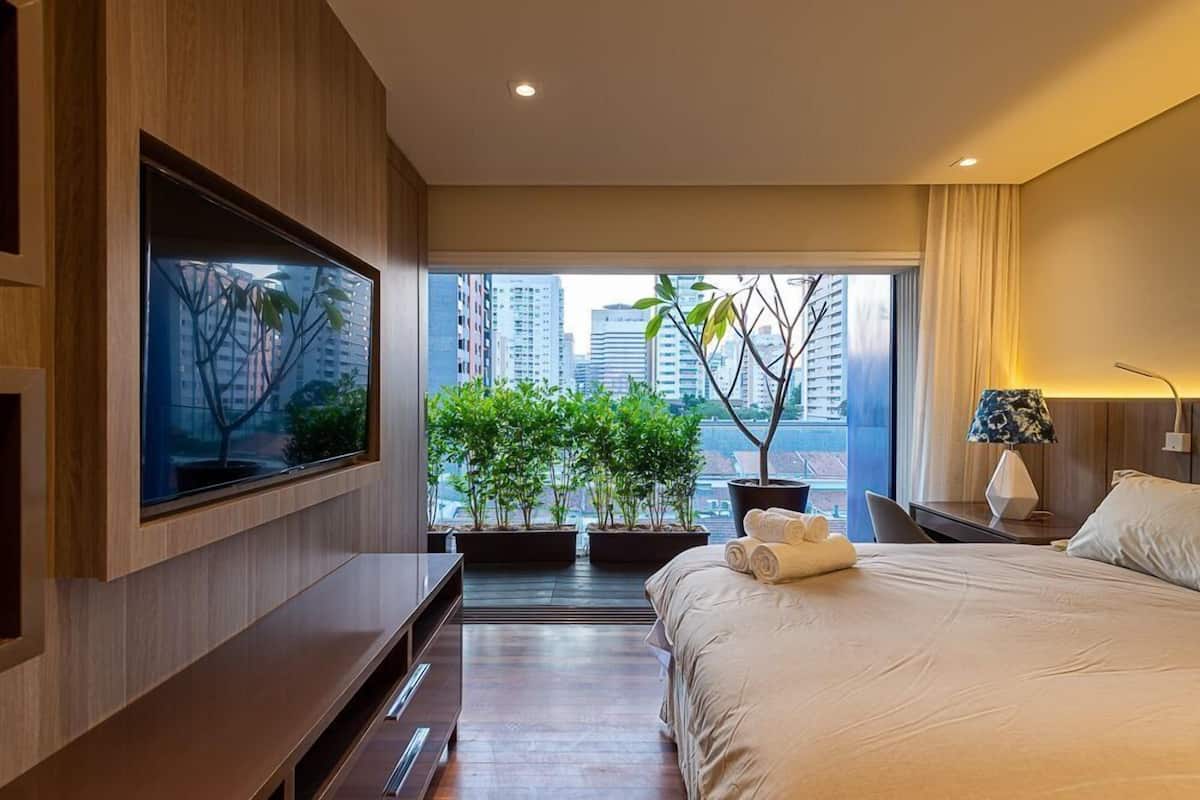 Ein modernes Hotelzimmer mit einem großen Bett, einem flachbildfernseher an der Wand, einem Schreibtisch mit Lampe und Blick auf die Stadt durch große Fenster.