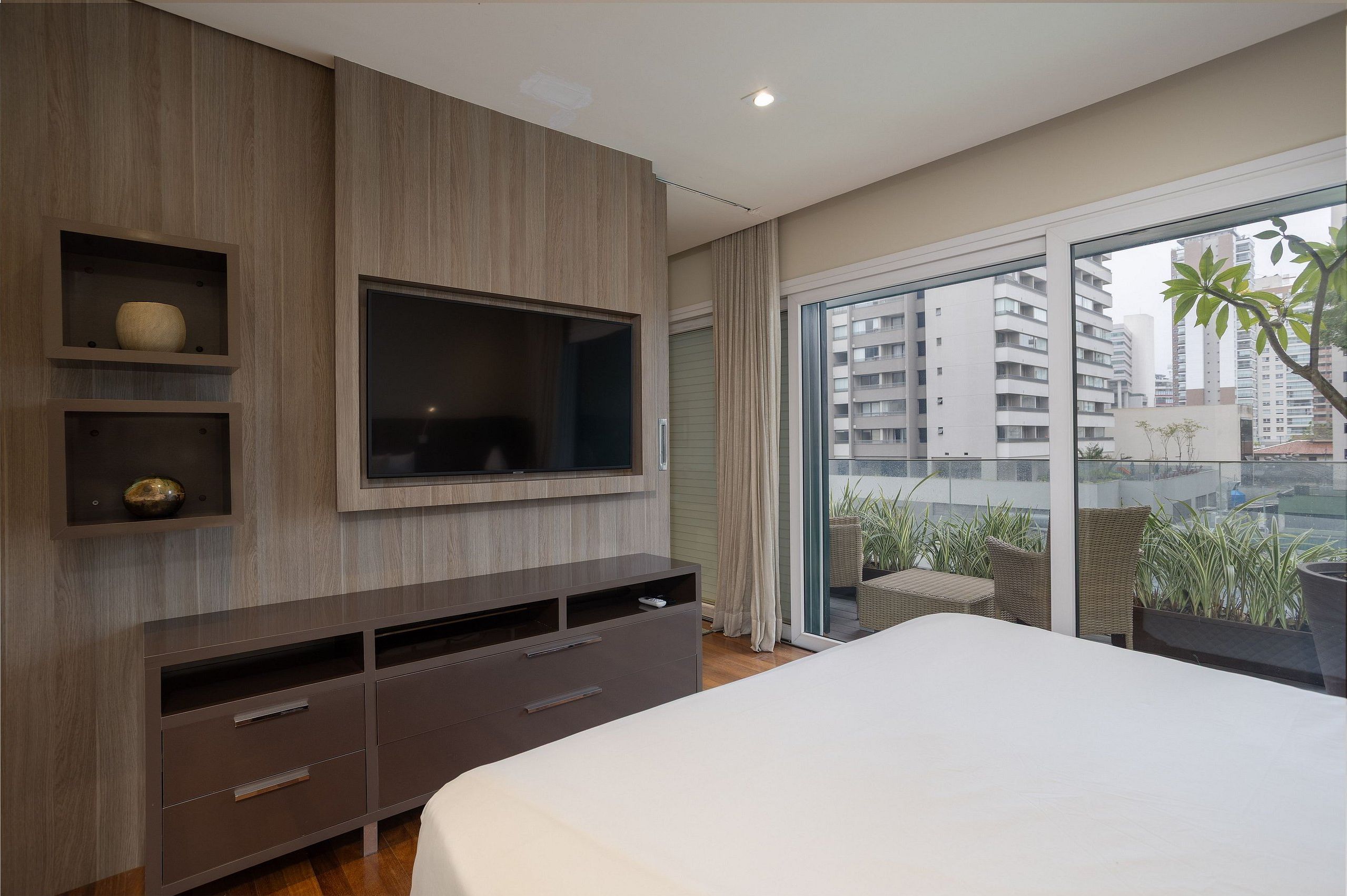 A modern bedroom with a large bed, a TV mounted on the wall, and a balcony with wicker furniture.