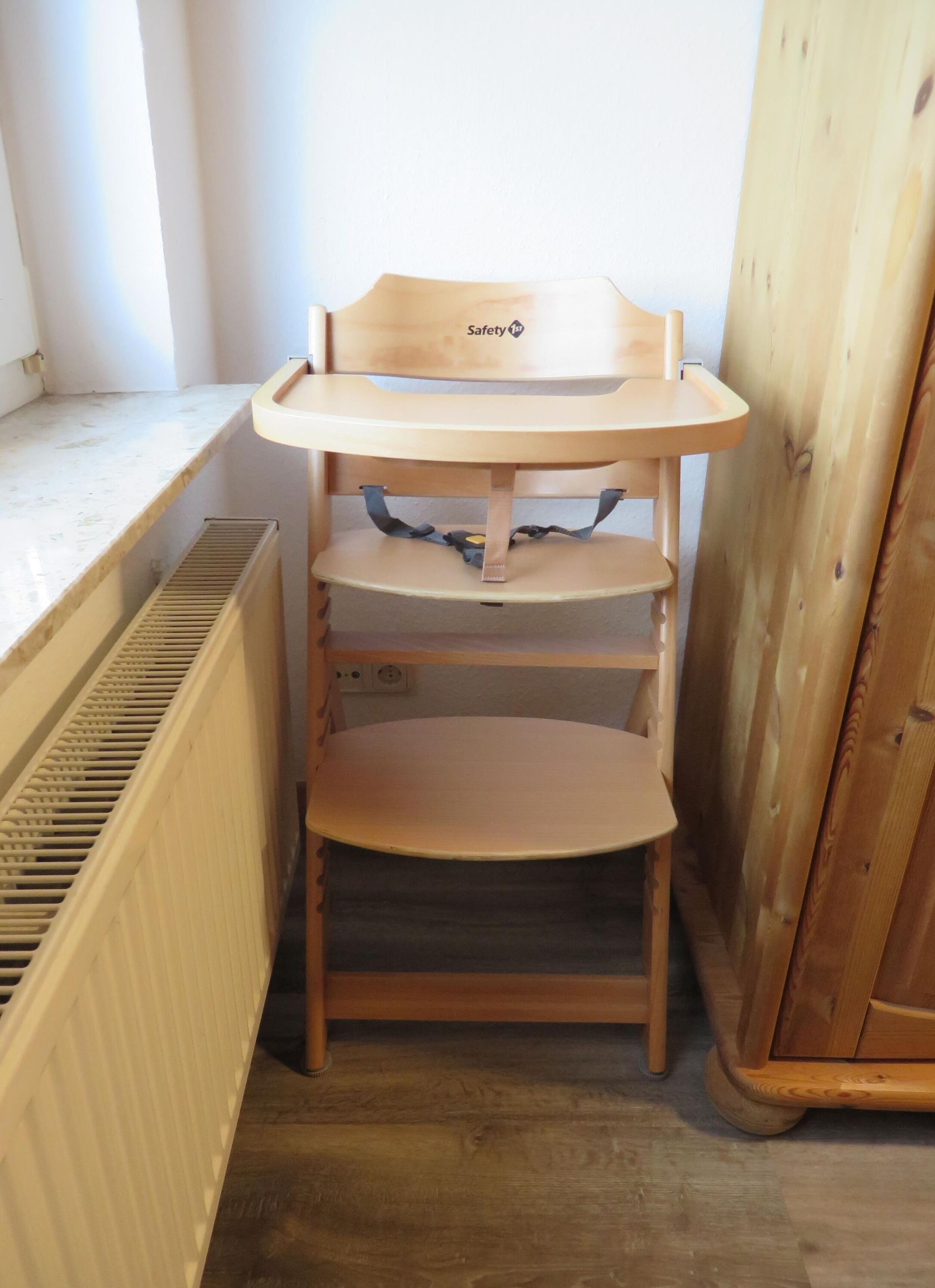 Une chaise haute en bois, munie d’une étiquette de sécurité, est placée près d’une fenêtre, au-dessus d’un radiateur.