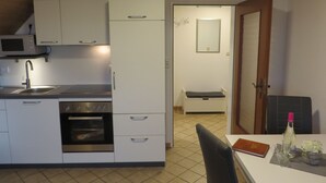 Une cuisine compacte avec des meubles blancs, un four encastré et un micro-ondes. On y trouve également un coin repas avec une table, des chaises et une bouteille de vin.