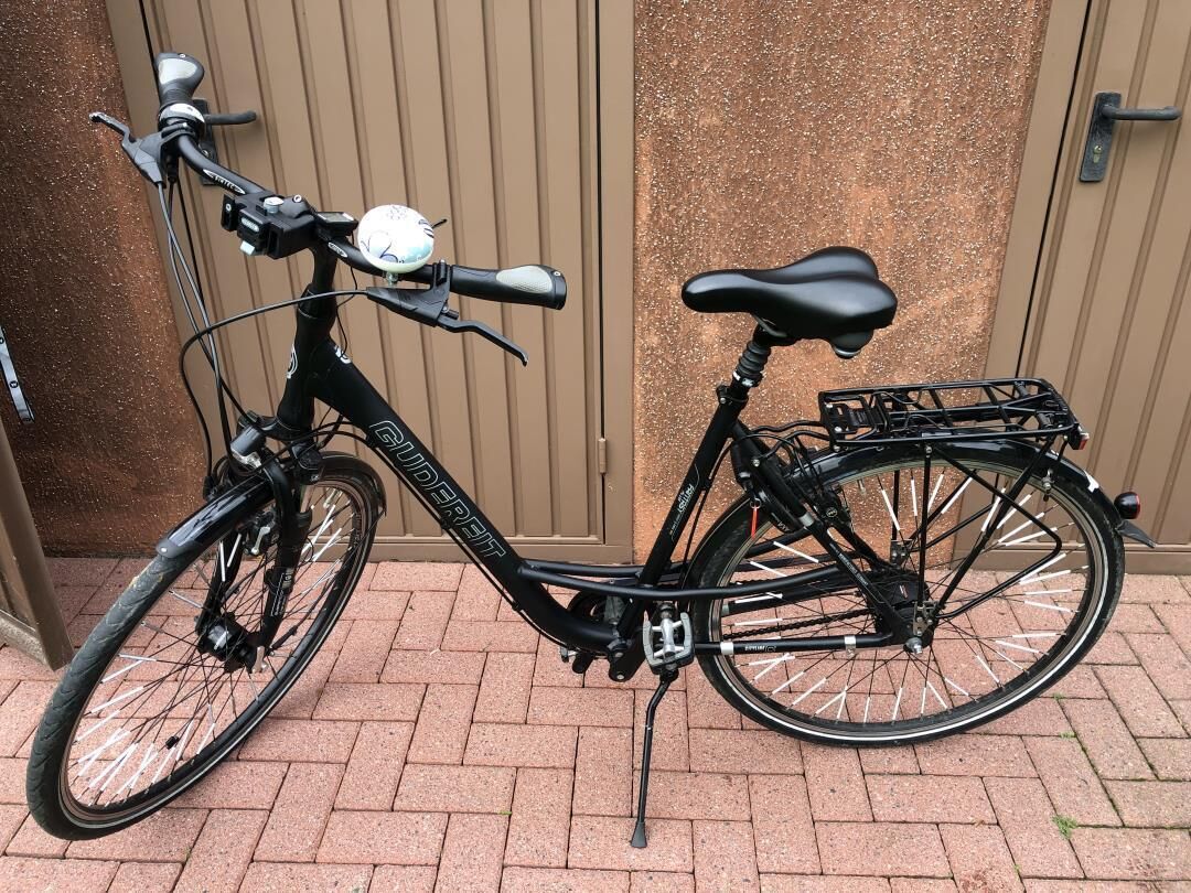 A stationary black bicycle with a basket, parked on a brick surface.