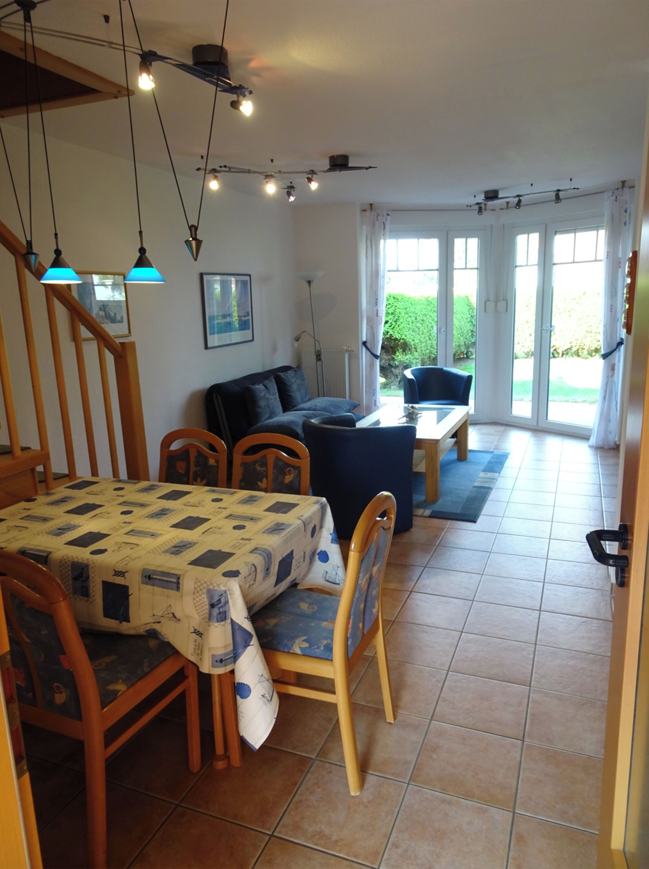 Zona pranzo con tavolo imbandito per quattro persone, una poltrona e una sedia. Luci a soffitto e vista sull'esterno tramite una porta vetrata.