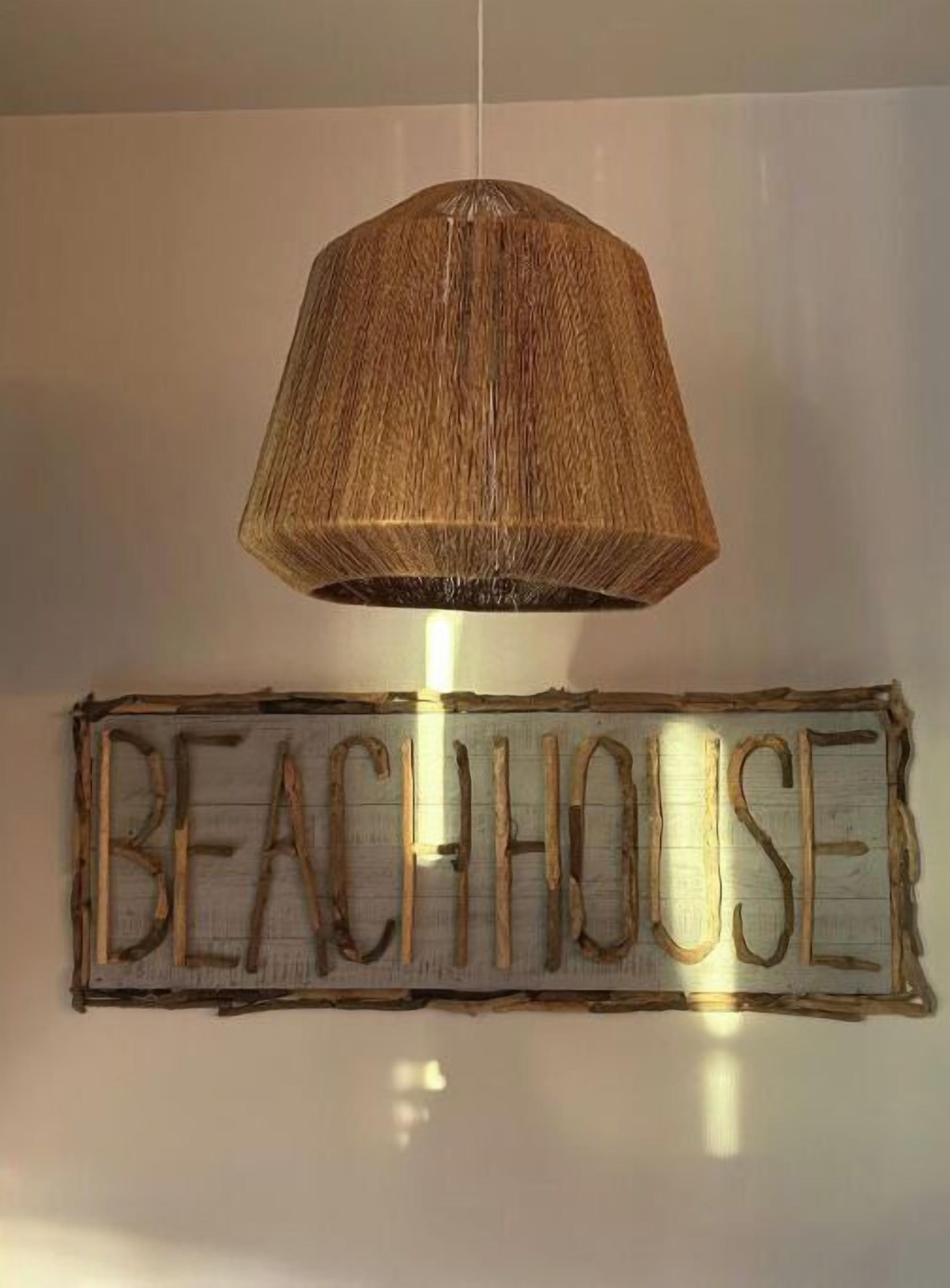 Een hanglamp met een gevlochten kap hangt boven een rustiek bord waarop 'BEACHHOUSE' staat.