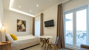 A modern hotel room with a bed, a small table, a TV, and a view of the outside. - Baia Flegrea Meublè - Deluxe Suite with terrace (Bacoli)