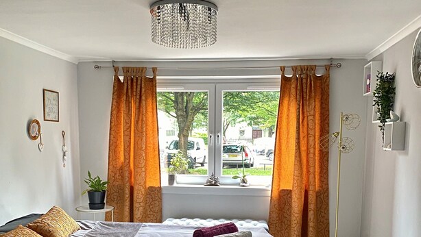 A hotel room with a bed, two rolled towels, a chandelier, a window with curtains, a potted plant, and a floor lamp. - Apartment Sleeps 5 With Free Parking (Edinburgh)
