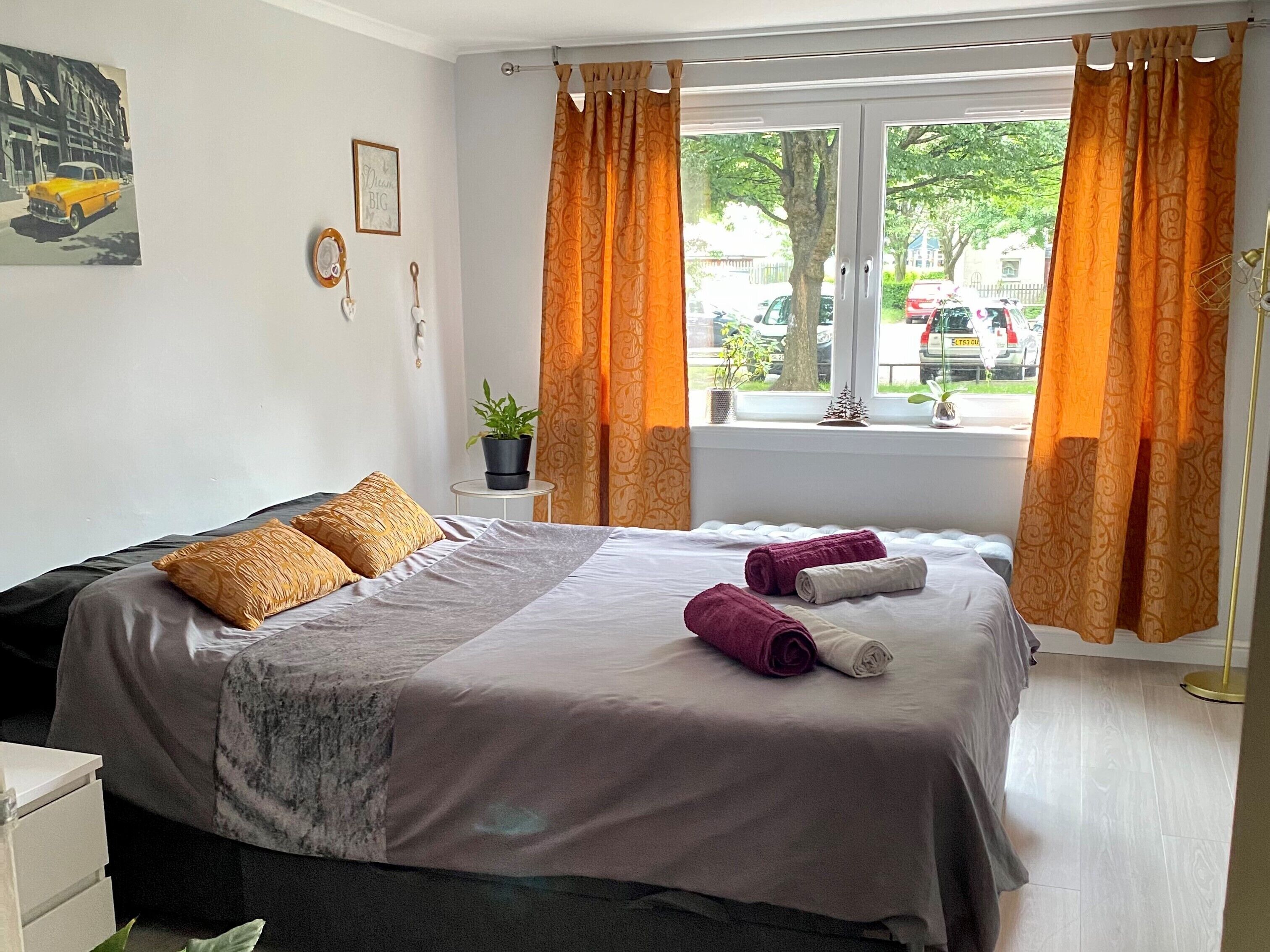 A hotel room with a bed, a window with curtains, a small table with a plant, and a wall with a picture.
