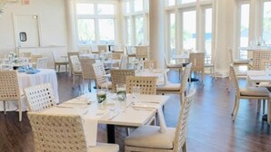 A spacious dining area with high ceilings and large windows, featuring white wicker furniture and a polished wooden floor.