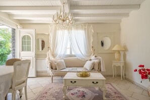 Ein Zimmer mit einem weißen Sofa, einem weißen Tisch, einer Kristallleuchter, einer Lampe und einer Vase mit roten Blumen.