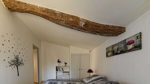 Une chambre avec un grand plafond en bois apparent, un lit avec une literie à motifs, une bibliothèque et un mur orné d’un tableau décoratif.