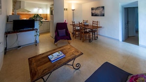 Un salon avec un téléviseur, une table basse en bois, un canapé, une table à manger avec des chaises, et une cuisine en arrière-plan.