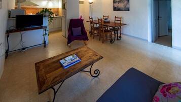 Un salon avec un téléviseur, une table basse en bois, un canapé, une table à manger avec des chaises, et une cuisine en arrière-plan.