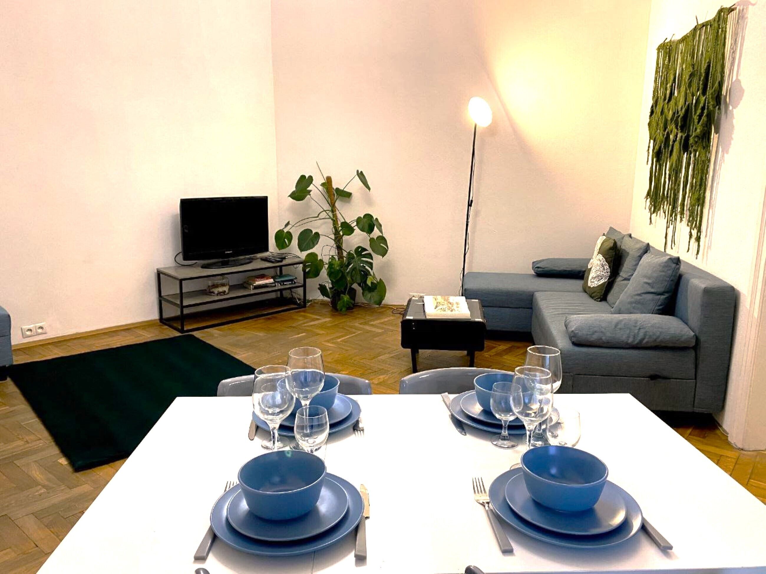 Un coin repas avec une table blanche dressée pour deux personnes, un canapé gris, un téléviseur posé sur un meuble et une plante.