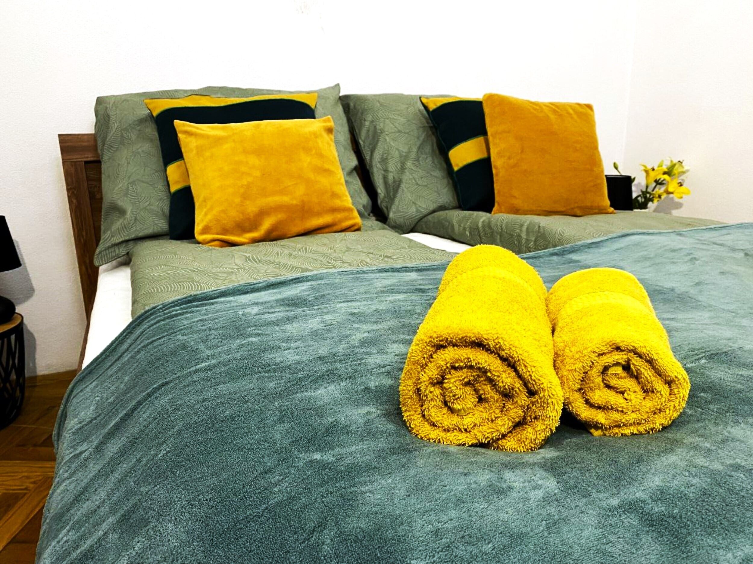A neatly made bed with a green blanket, two pillows, and two rolled towels.