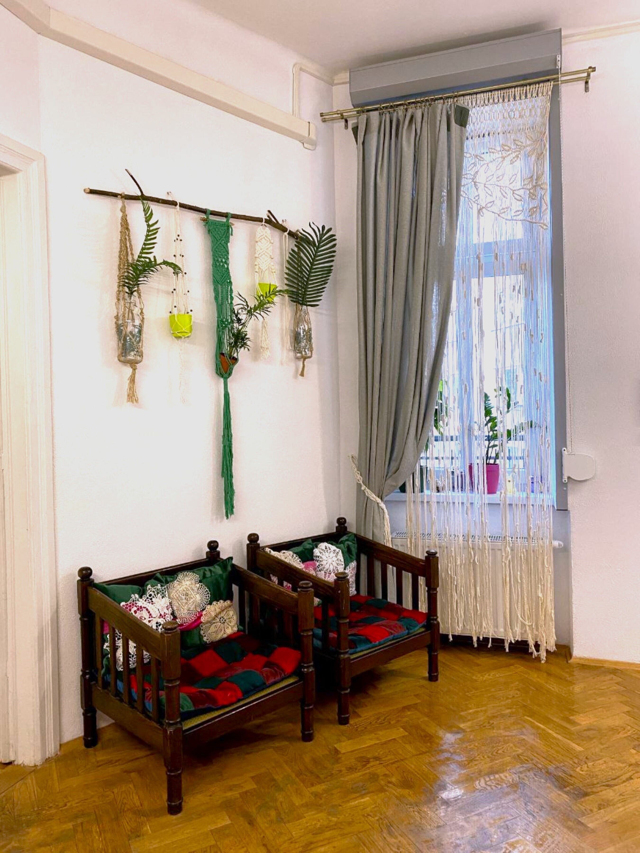 Quarto com duas cadeiras de madeira, piso de madeira, janela com cortina e decorações nas paredes, incluindo plantas pendentes e macramé.