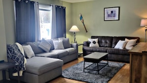 Ein Wohnzimmer mit grauem Ecksofa, einem zusätzlichen Sofa, einem Couchtisch und einer Lampe. Ein Fenster mit Vorhängen, ein Bild an der Wand und ein Holzboden sind ebenfalls vorhanden.