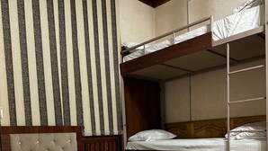 Une chambre d’hôtel avec un lit simple, un lit superposé, une tête de lit en bois, une table de chevet avec un verre, une télécommande et un petit appareil.