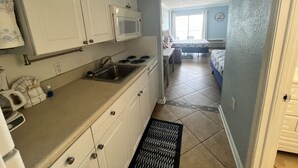 Een kleine keuken met witte kasten, een magnetron en een spoelbak. Een zwarte mat ligt voor de spoelbak en een waterkoker staat op het aanrecht.
