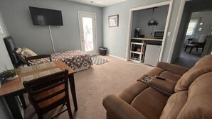 Ein Wohnzimmer mit braunem Sofa, einem Esstisch mit Stühlen, einem an der Wand befestigten Fernseher und einer Küche im Hintergrund.