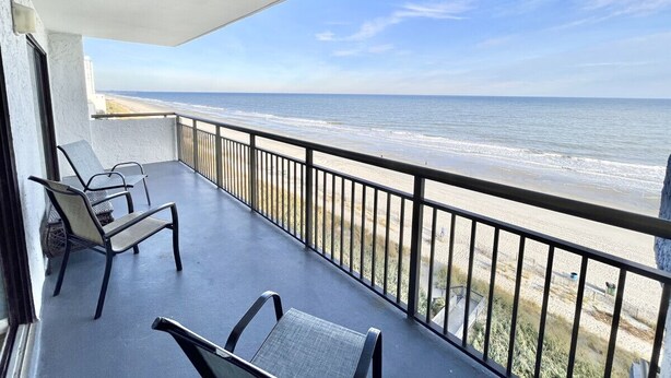 A balcony with two chairs and a table, overlooking a beach with a railing. - Stunning Oceanfront 2bd/2Ba Condo, Myrtle Beach (Myrtle Beach)