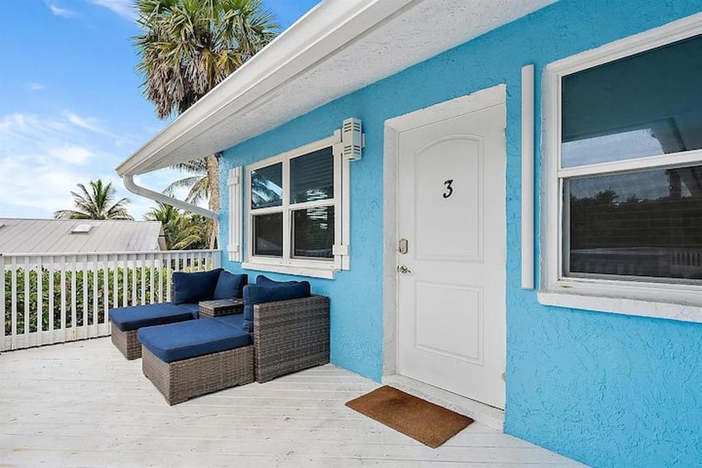 Une maison à l’extérieur bleu, avec une porte blanche portant le numéro 3, et un porche meublé de meubles en osier.