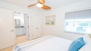 Une chambre à coucher comprenant un lit, un ventilateur de plafond, une fenêtre avec des stores et une porte donnant sur une salle de bain.