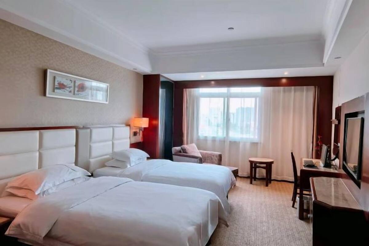 Une chambre d’hôtel avec deux lits, un bureau, une chaise, une fenêtre avec des rideaux et un tableau encadré au mur.