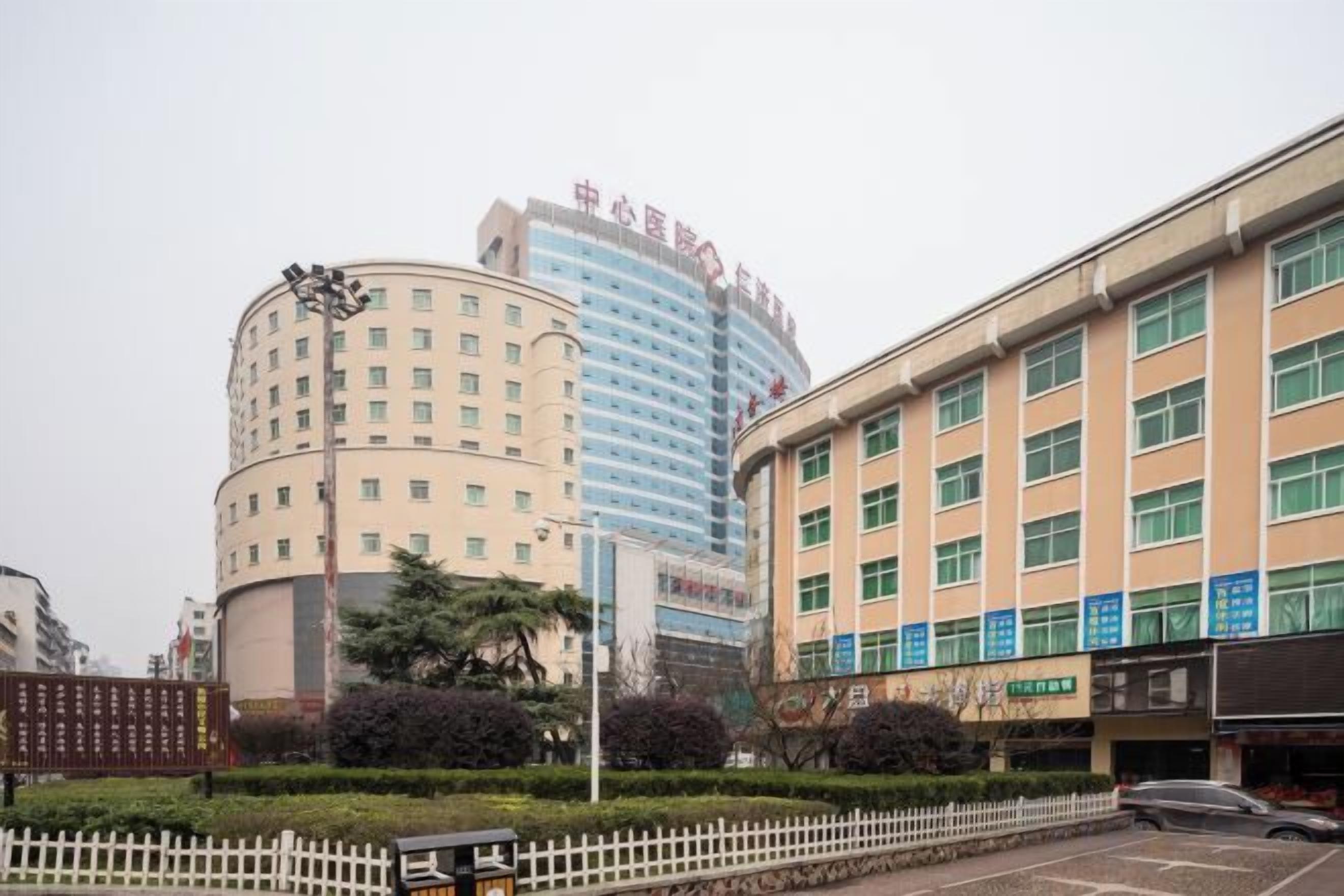 A multi-story hotel with a curved facade, a flat-roofed building adjacent to it, and a parking lot with a white fence.