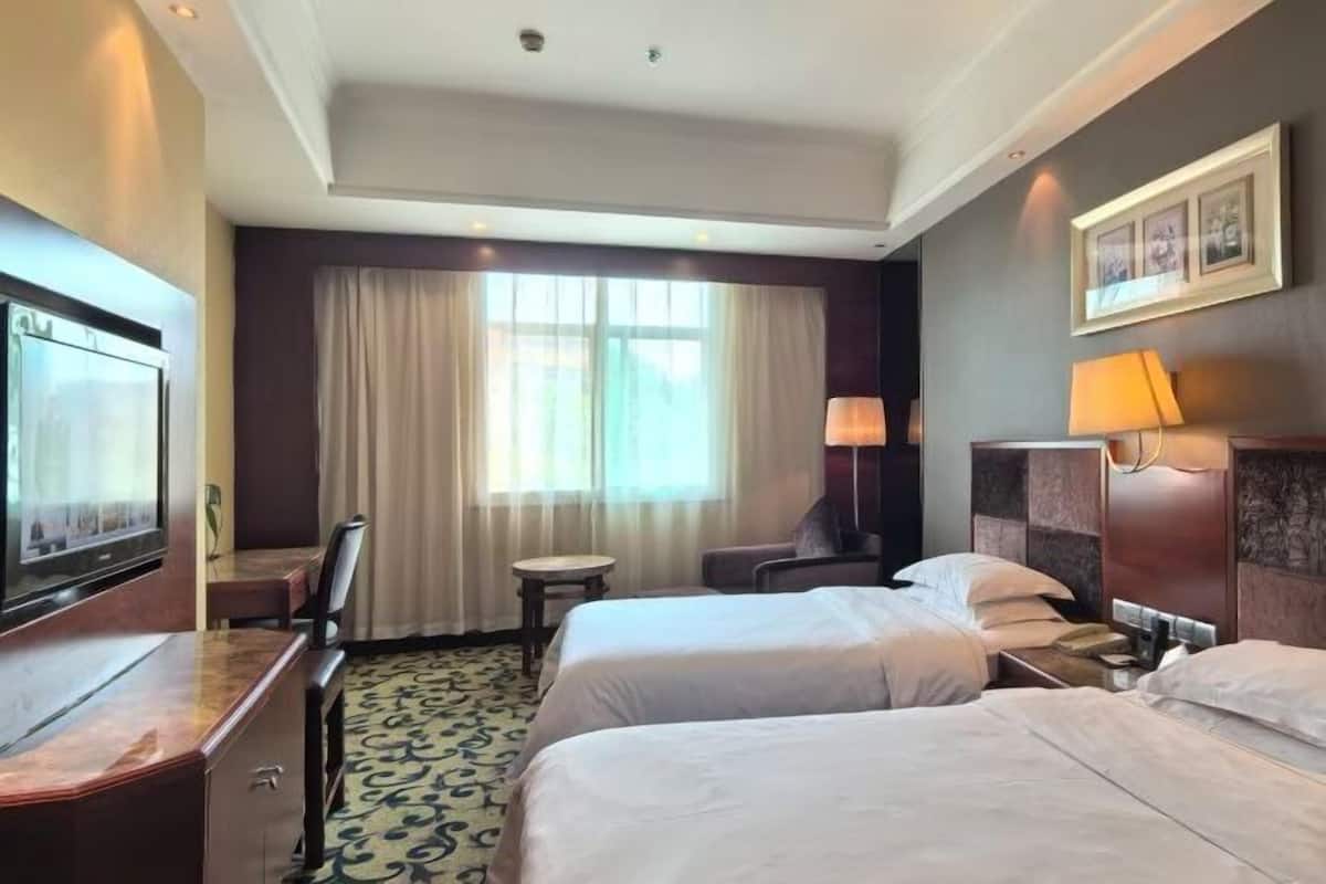 Une chambre d’hôtel avec deux lits, un bureau, une chaise, une télévision, une lampe et une fenêtre avec des rideaux.