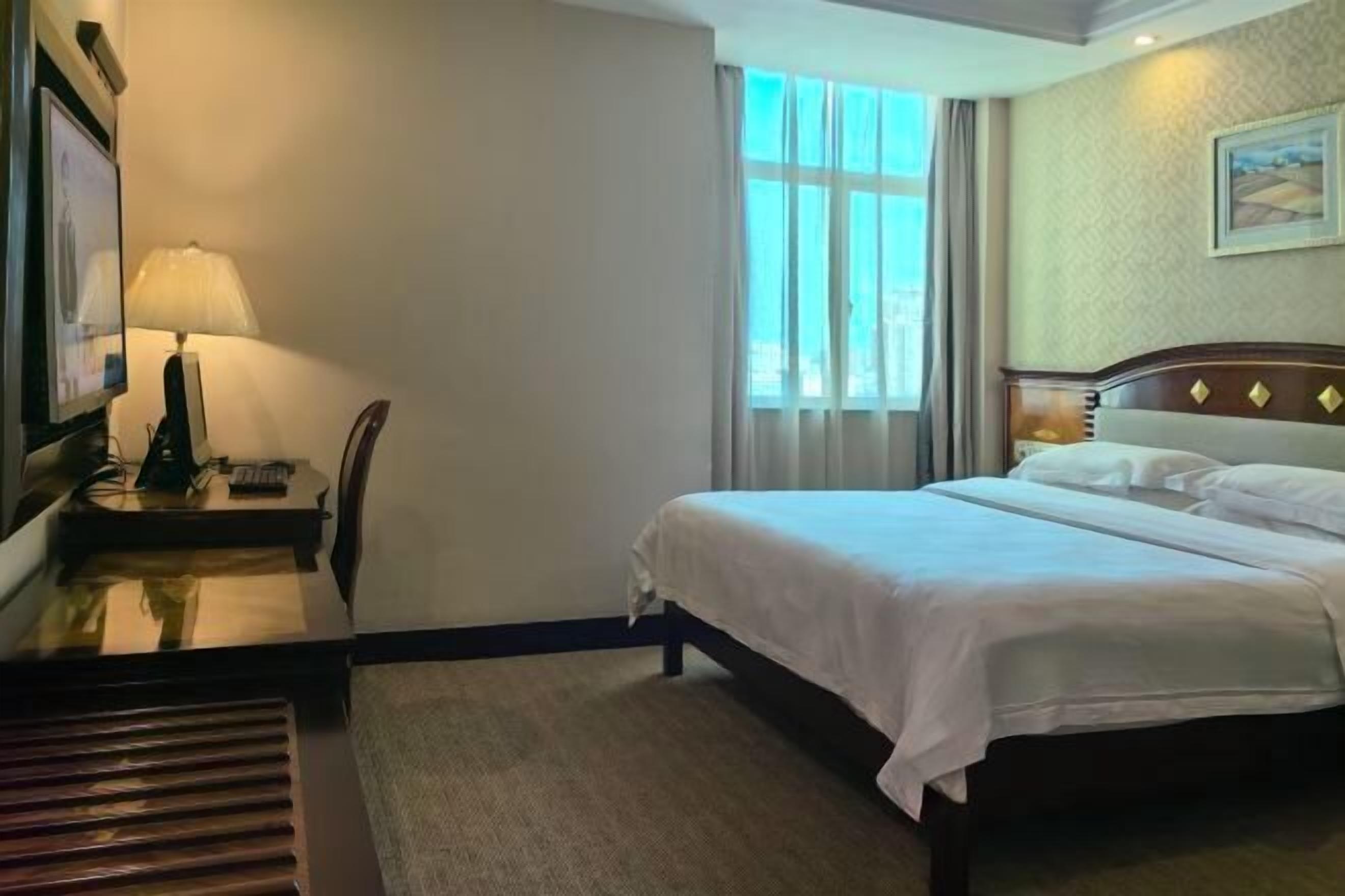 Habitación de hotel con cama, escritorio con lámpara, silla y ventana con cortinas.