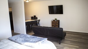 A bedroom with a bed, a black leather sofa, a desk with a computer, and a television on the wall.