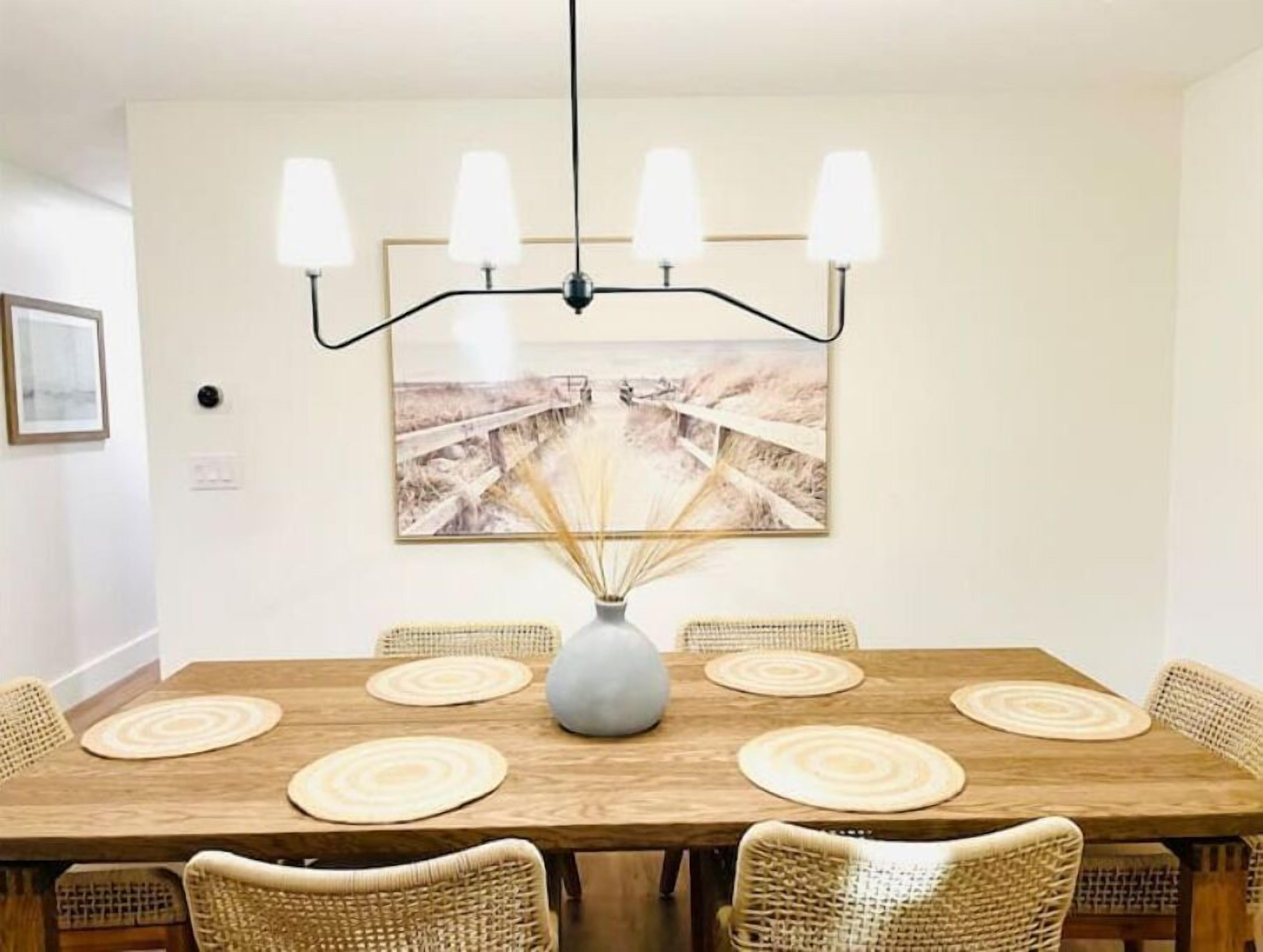 Un coin repas comprenant une table en bois, six chaises en osier, un vase central et un luminaire fixé au mur.