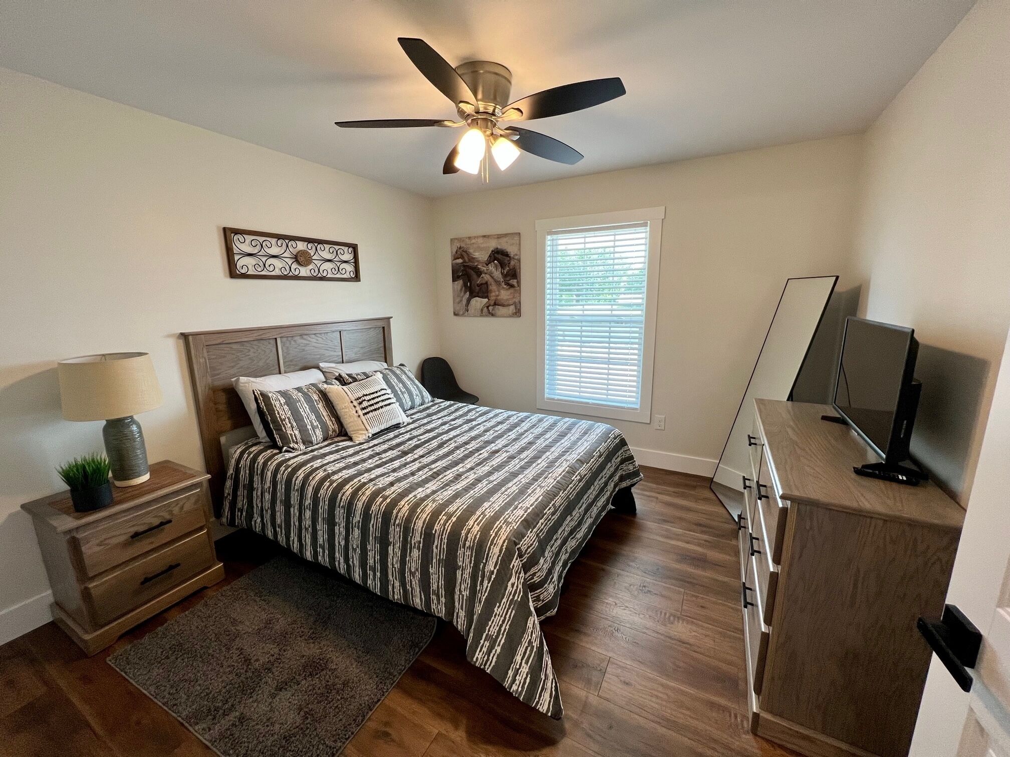 A bedroom with a bed, a nightstand, a lamp, a ceiling fan, a window with blinds, a TV on a stand, and a dresser.