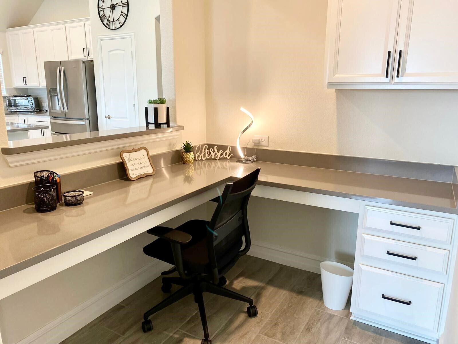 A modern kitchen with white cabinets, a built-in oven, a refrigerator, a sink, and a countertop with a chair and a trash bin.